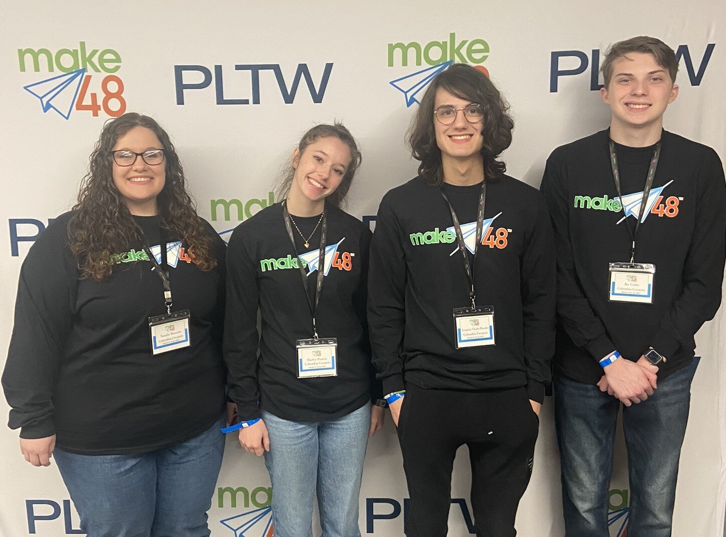 Three teenage students from the Columbia Creators team stand to the right of their teacher. All are wearing the same forest green, long-sleeved shirt and standing in front of a make48 and PLTW banner. 