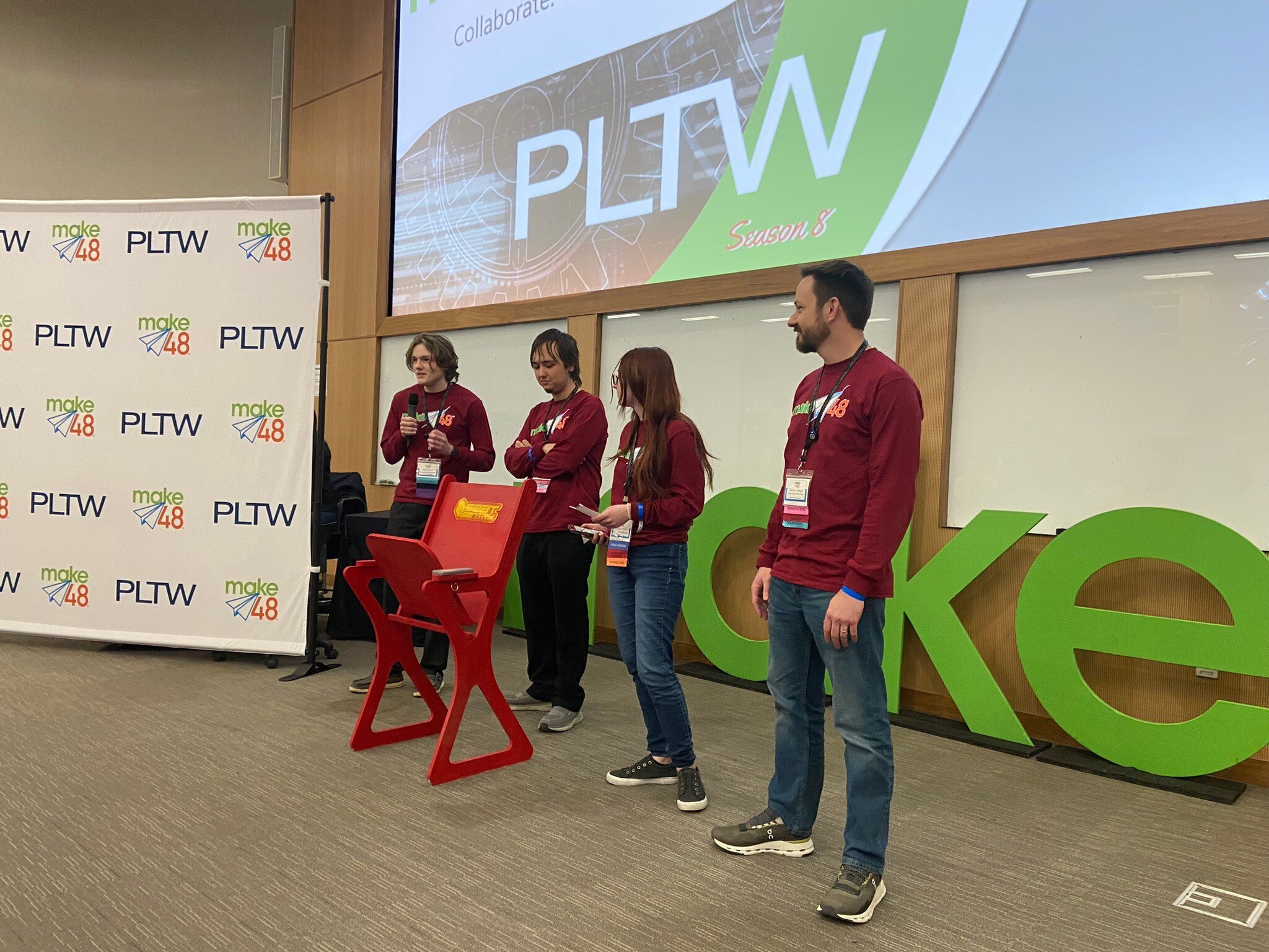 The Innovation Hall Bears team members and one of their teachers stands on stage with a red, folding stadium seat in front of them. They are presenting their prototype to the judges. 