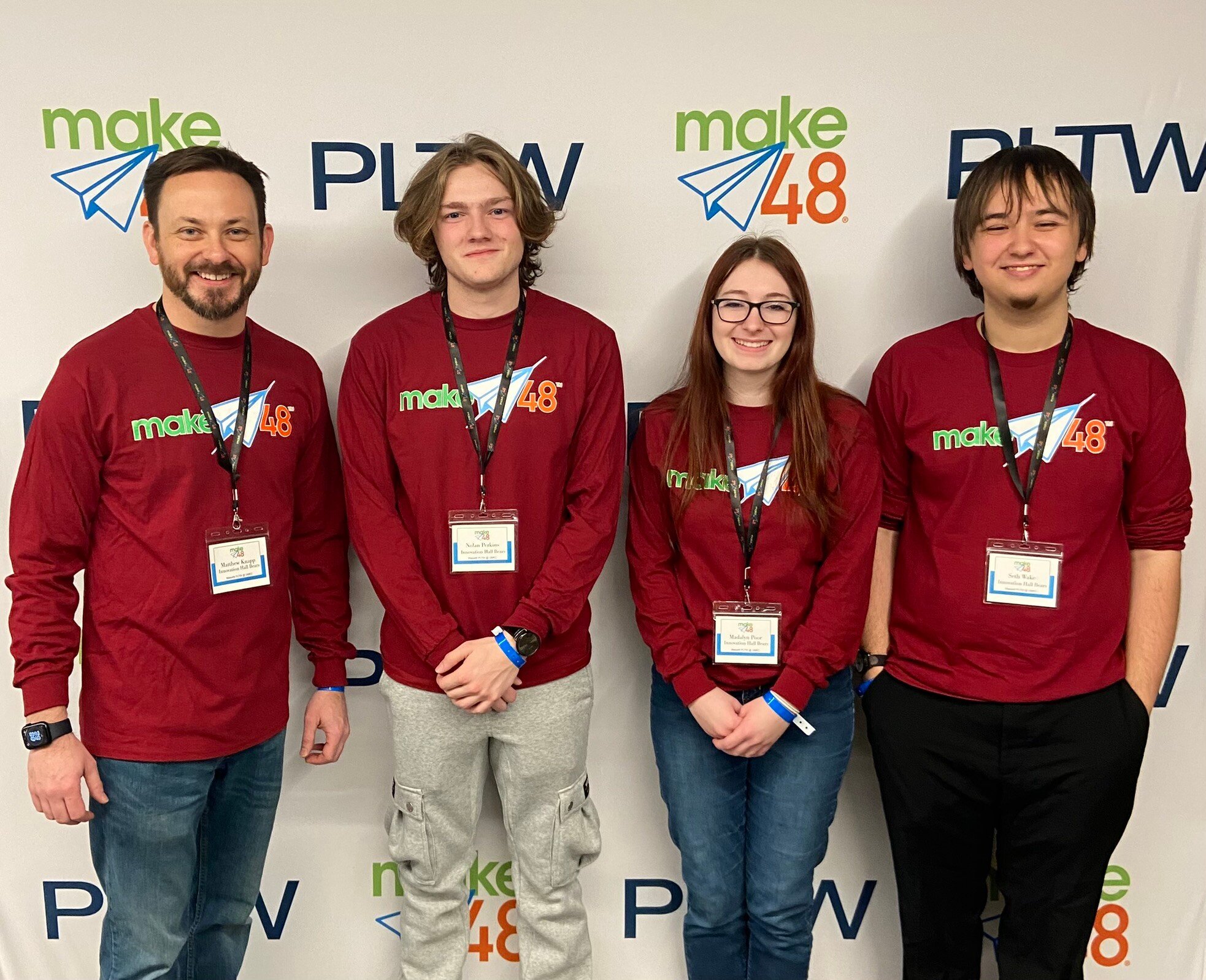 Three teenage students from the Innovation Hall Bears team stand to the right of their teacher for a group photo. All are wearing the same dark red, long-sleeved shirt and standing in front of a make48 and PLTW banner. 