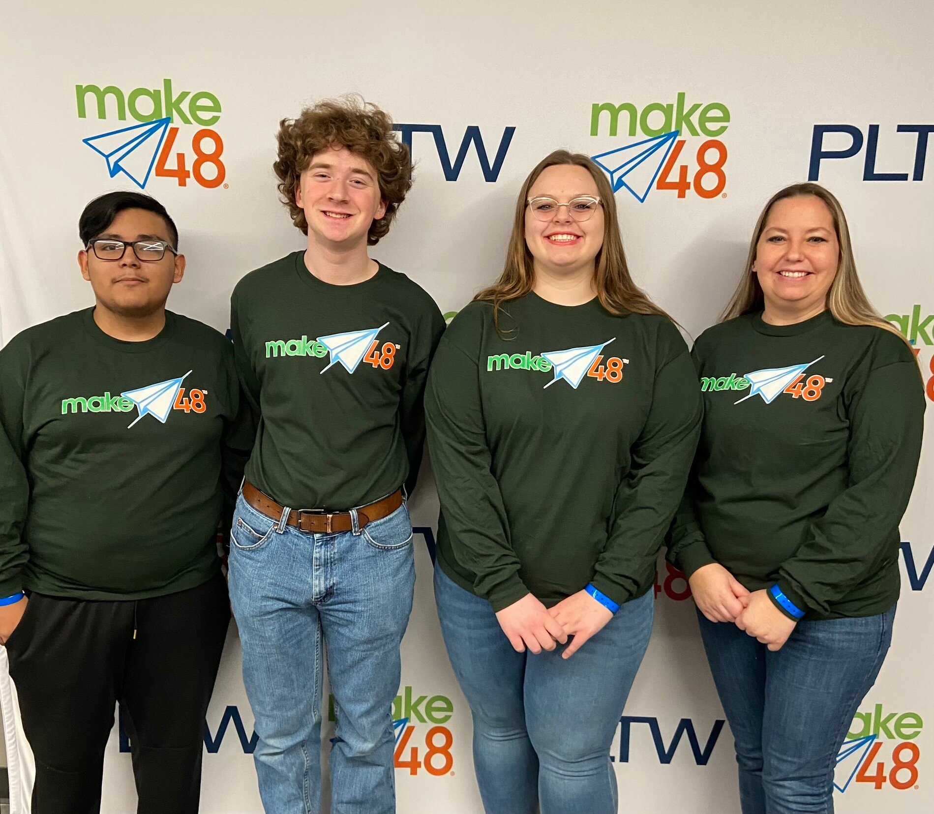 Three teenage students from The Ones Who Shall Not Be Named team stand to the Left of their teacher for a group photo. All are wearing the same tan, long-sleeved shirt and standing in front of a make48 and PLTW banner. 