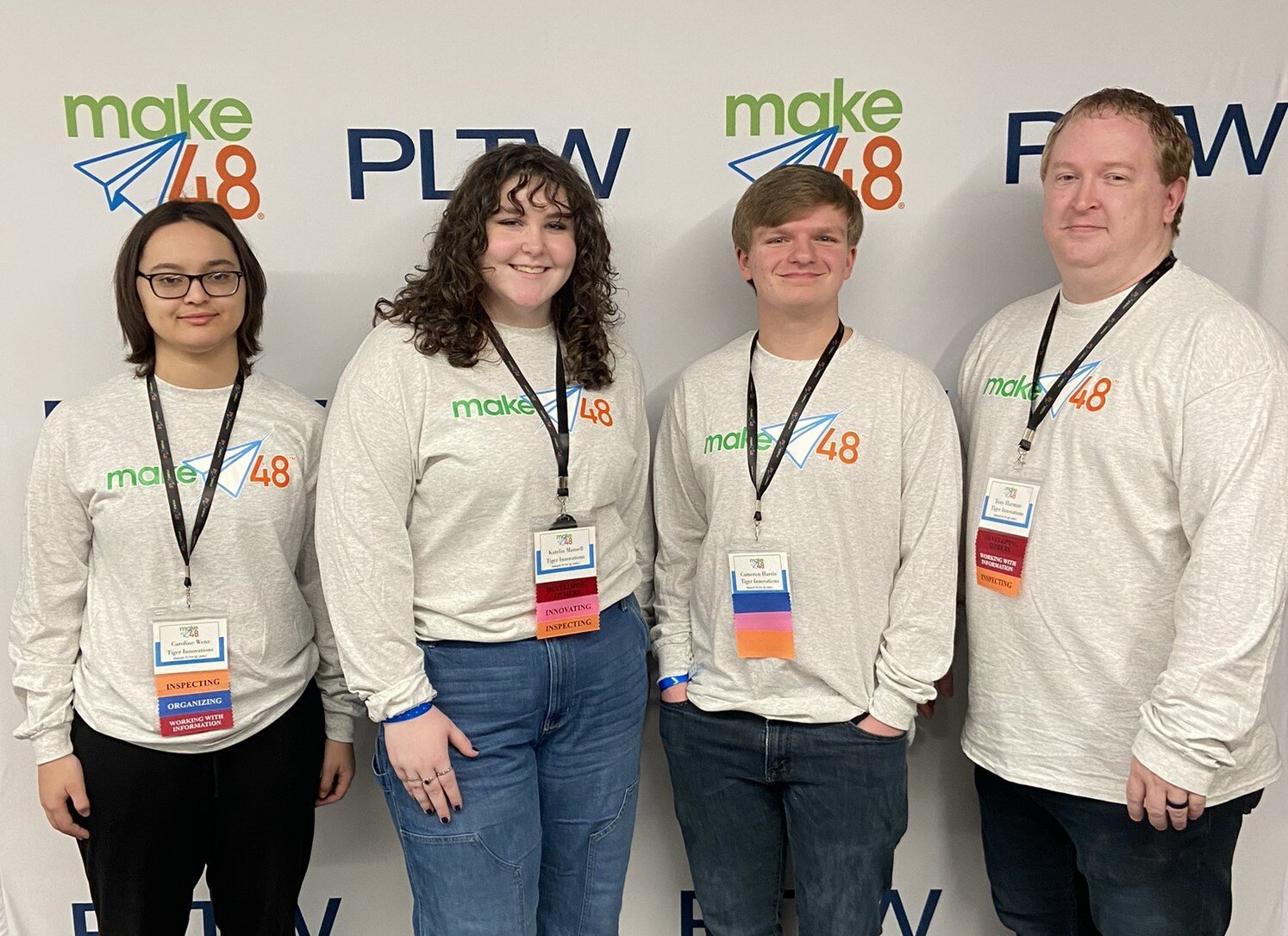 Three teenage students from Tiger Innovations team stand to the Left of their teacher for a group photo. All are wearing the same tan, long-sleeved shirt and standing in front of a make48 and PLTW banner. 