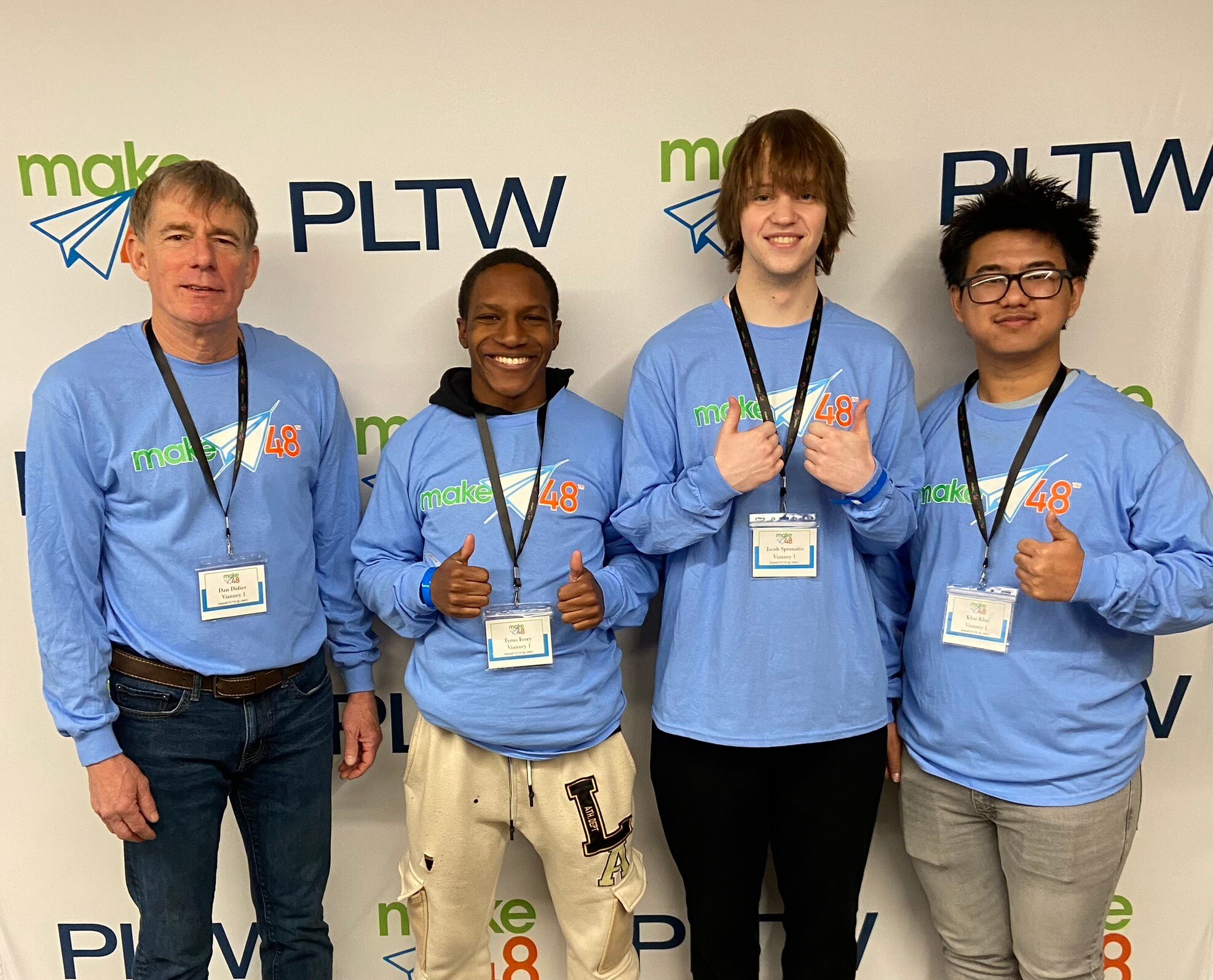 Three teenage students from the Vianney 1 team stand to the right of their teacher for a group photo. All are wearing the same light blue, long-sleeved shirt and standing in front of a make48 and PLTW banner. 